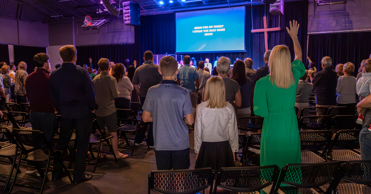 Congregation standing and worshiping with a woman in a green dress raising her hand in a church setting with a cross and blue screen displaying worship lyrics.