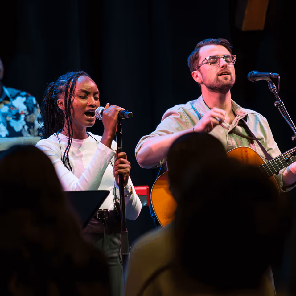 Female singer with braided hair singing into a microphone beside a male guitarist wearing glasses on stage during a live performance.