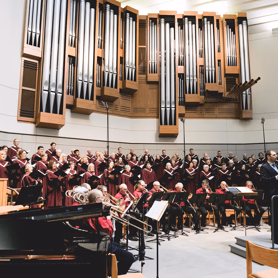 Choir in maroon robes singing with orchestra musicians playing brass instruments and a conductor leading in front, beneath large organ pipes.