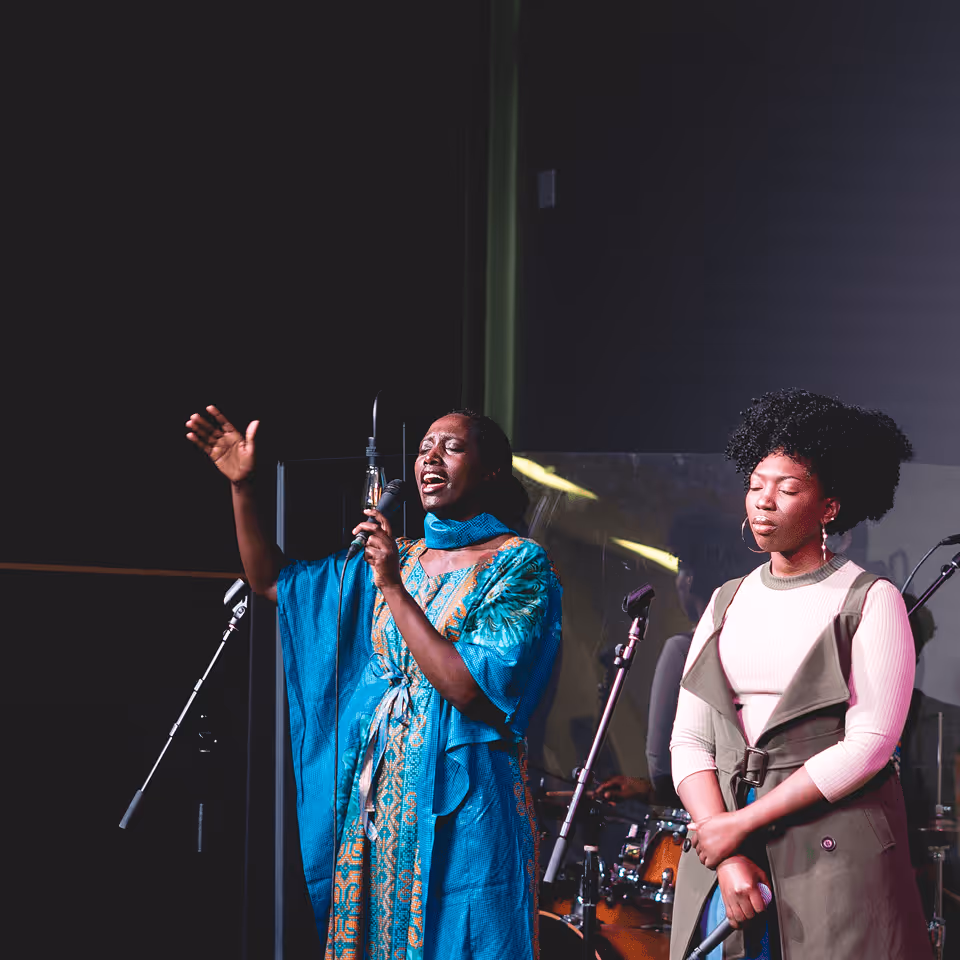 Two women performing on stage; one passionately singing into a microphone wearing a blue patterned dress, the other with eyes closed holding a microphone, wearing a white top and green vest.