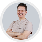 Man with short dark hair wearing a light beige shirt and smiling with arms crossed.