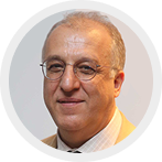 Portrait of a middle-aged man with short gray hair, glasses, and a slight smile, wearing a white collared shirt and a dark blazer.