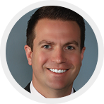 Smiling man with short dark hair wearing a dark suit and white shirt against a gray background.