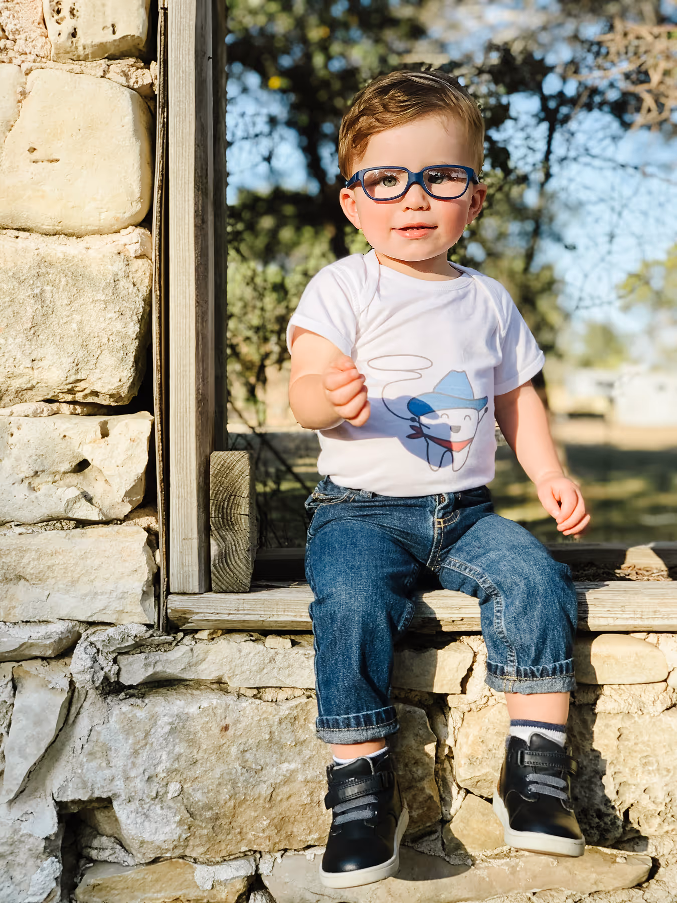A little boy sitting on a window sill wearing glasses.