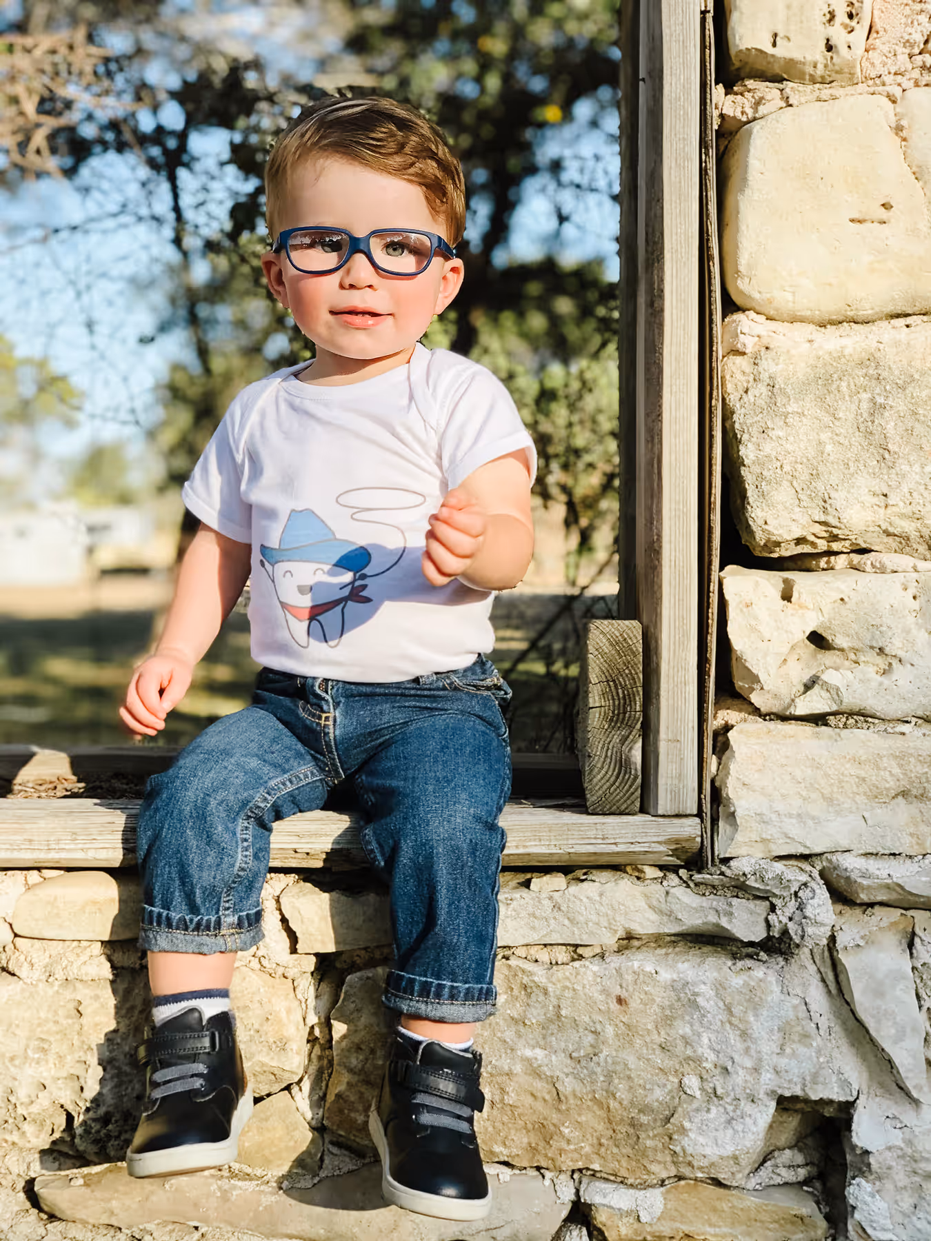 A little boy wearing glasses sitting on a window sill.