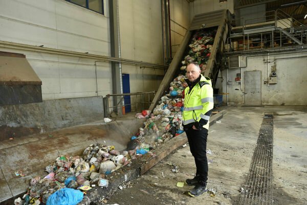 En mann i gul high-visibility jakke står inne på et avfallsmottak ved siden av et transportbånd fylt med søppel.
