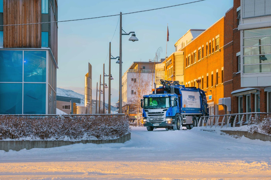 Blå renovasjonsbil kjører på snødekt vei mellom moderne bygninger i et vinterlandskap.