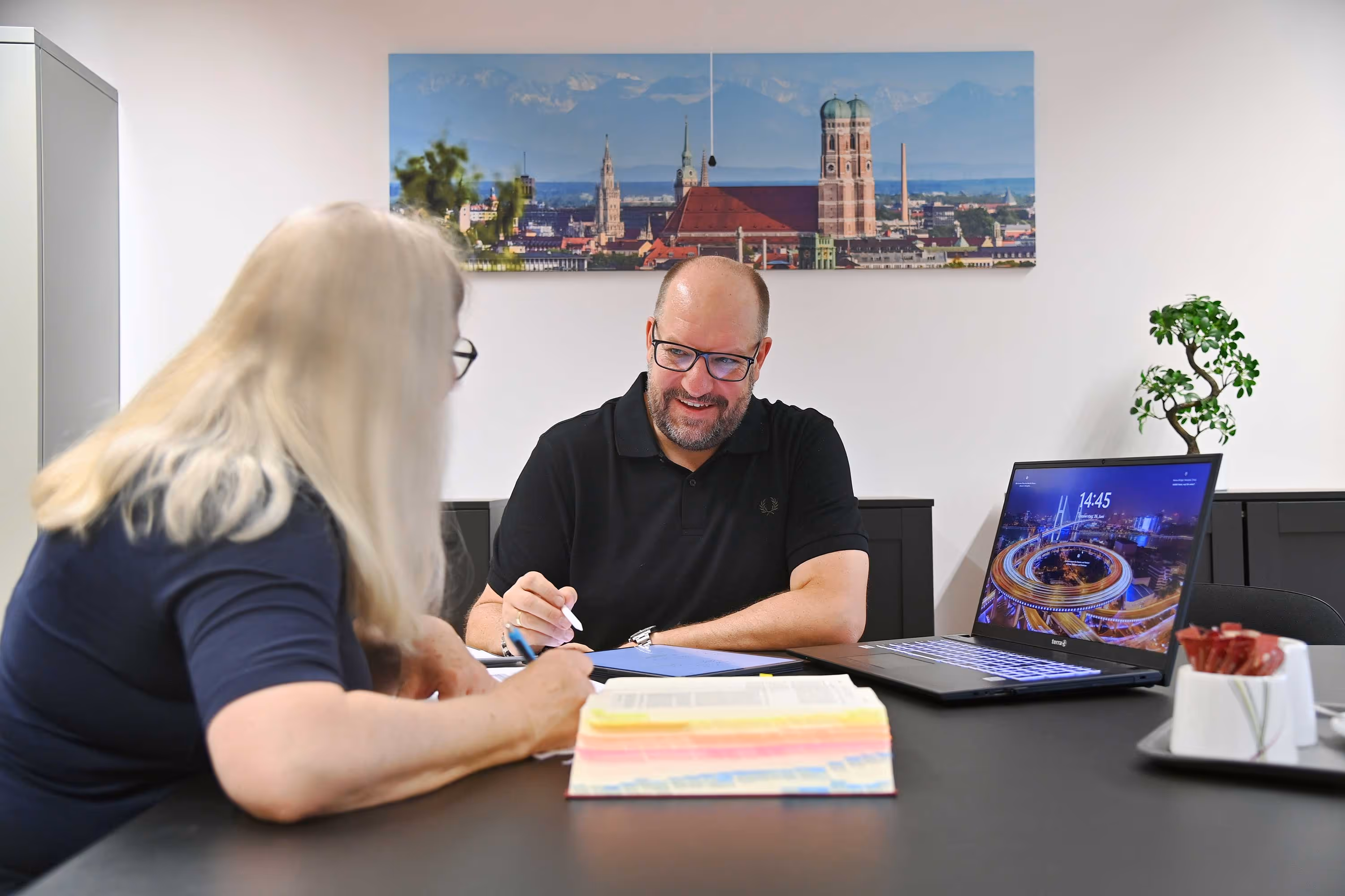 Seven people sitting on various colored scooters and a motorcycle in a paved courtyard with a white building and trees in the background.