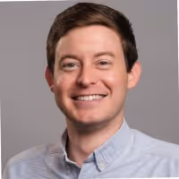 Smiling man with short brown hair wearing a light blue button-up shirt against a gray background.