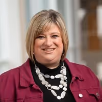 Smiling woman with short blonde hair wearing a maroon jacket and a white beaded necklace.