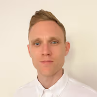 Portrait of a man with light skin and short blond hair, wearing a white collared shirt, against a plain light background.