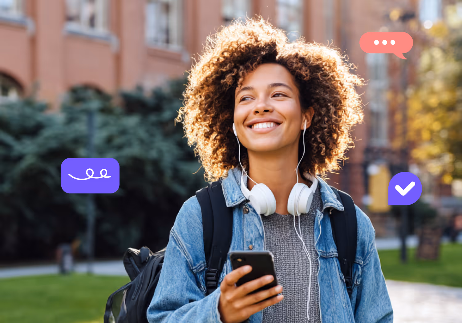 Smiling young woman with curly hair wearing a denim jacket and headphones around her neck holding a smartphone outdoors.