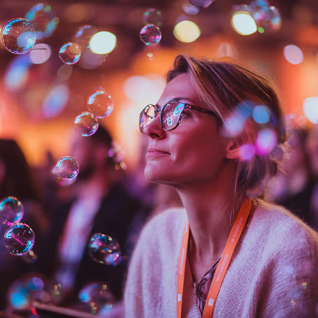 Woman wearing glasses and a conference badge looks ahead surrounded by floating bubbles in a dimly lit room.
