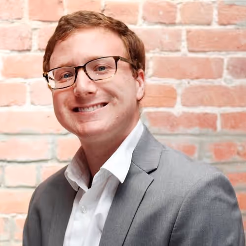 Smiling man with glasses wearing a grey suit and white shirt standing in front of a brick wall.
