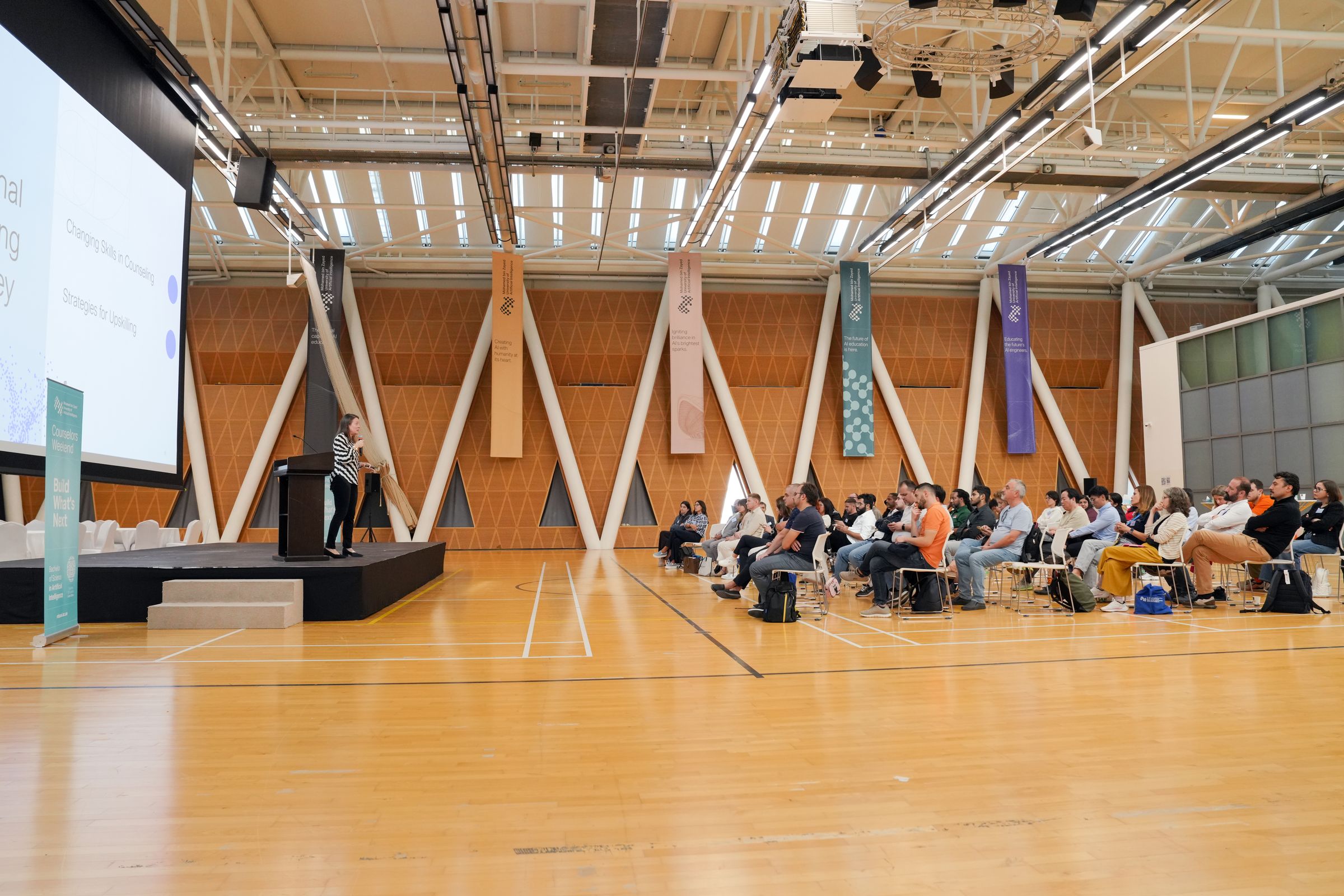 A speaker on stage in a large hall, with rows of attendees seated for the presentation