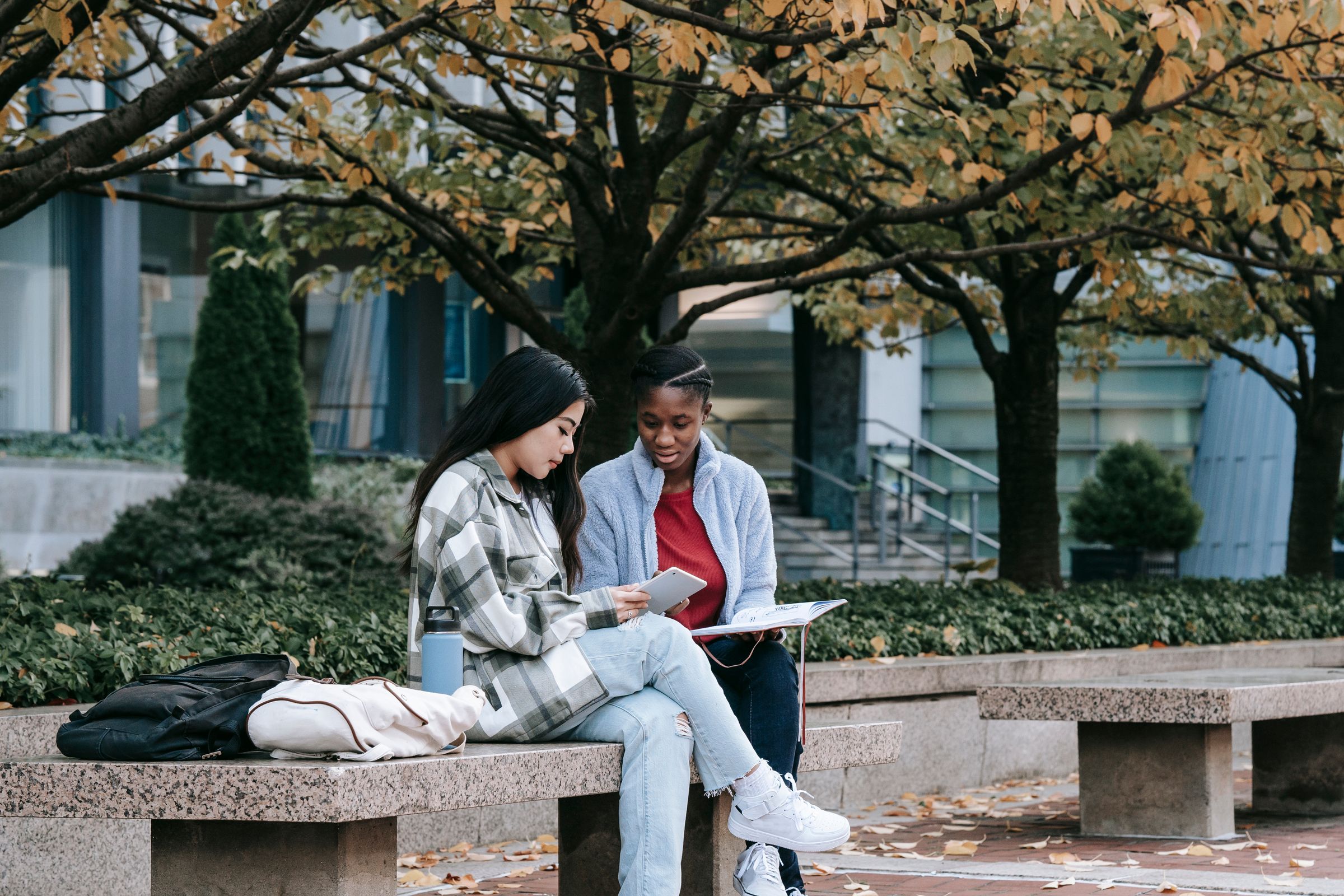 Two students sitting on a bench outside a campus building, looking at a tablet
