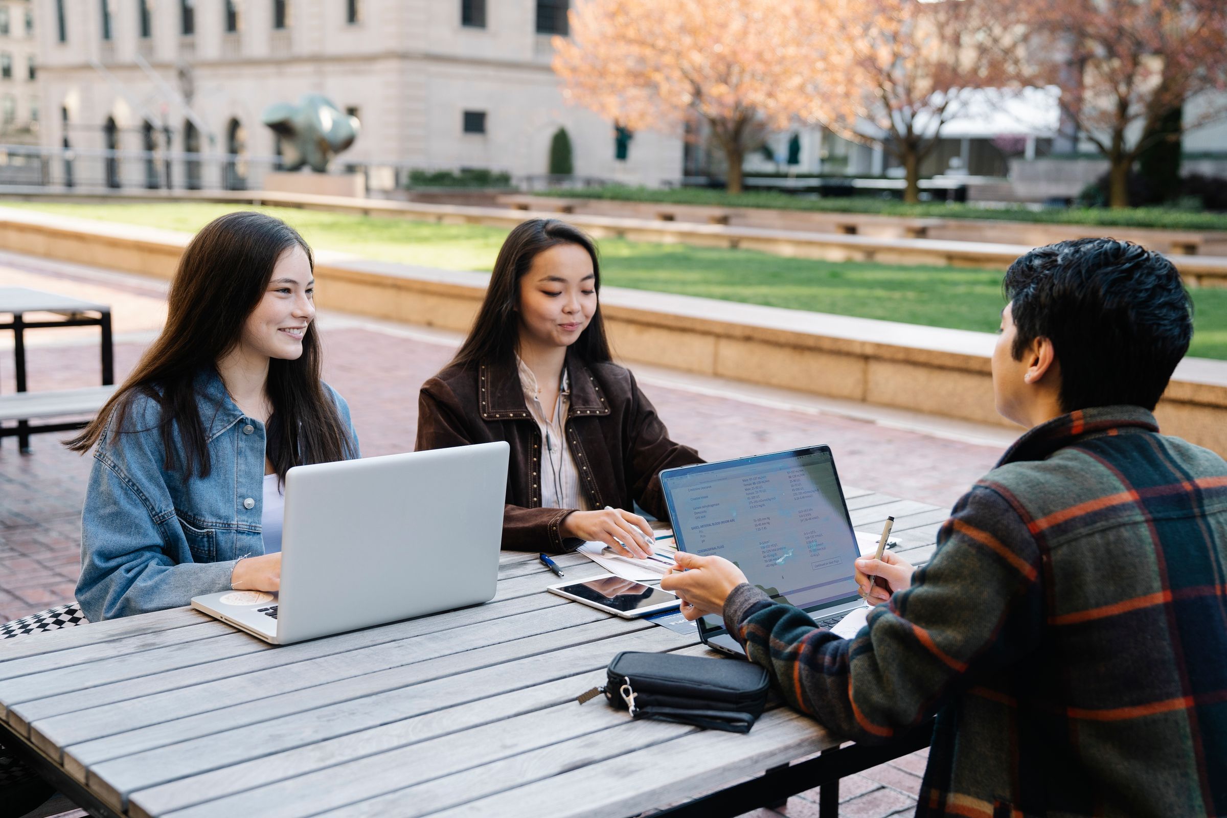 Three students working on laptops at an outdoor table on campus