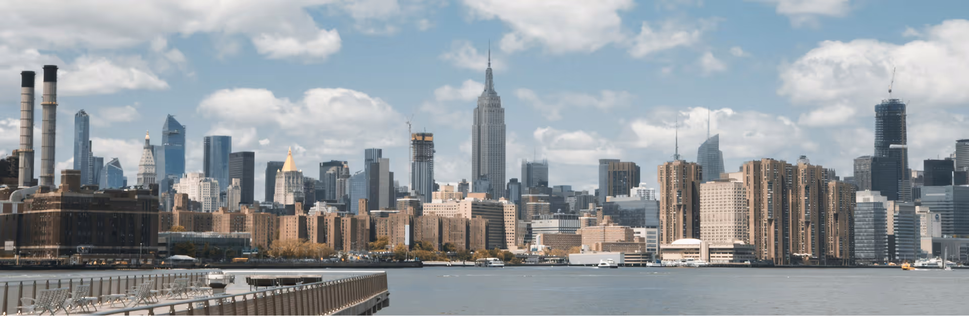 Skyline of a bustling city on a partly cloudy day. Prominent skyscrapers, including one tall, pointed building, rise above the waterfront, conveying urban energy.