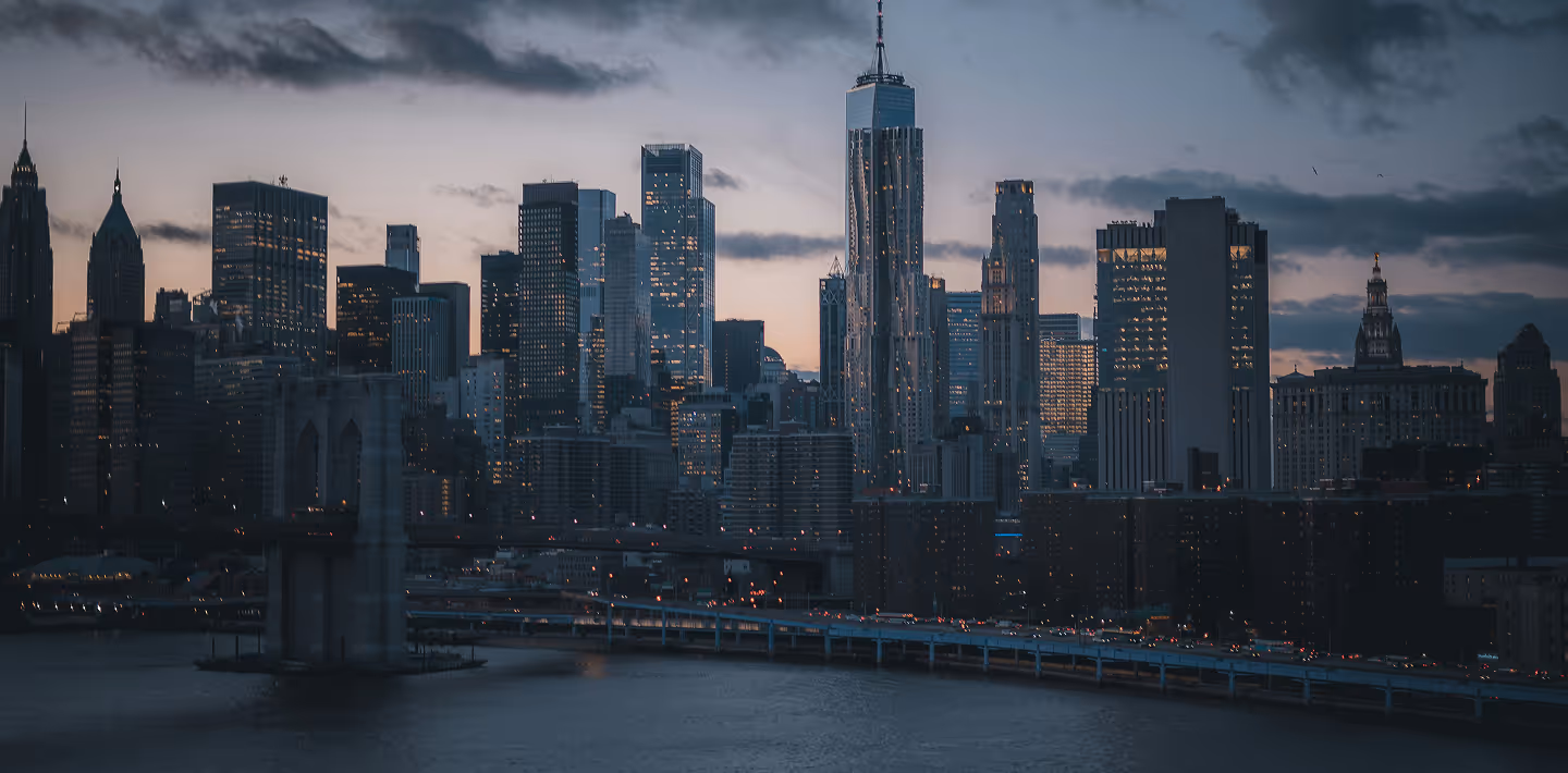 Twilight view of a city skyline with illuminated skyscrapers and a bridge in the foreground. The serene river reflects the city's lights, creating a calm ambiance.
