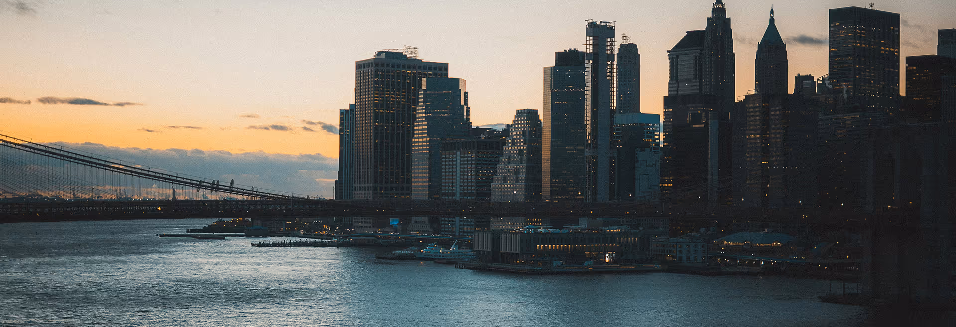 Skyline of a city at dusk with tall buildings silhouetted against an orange and blue sky. A bridge spans a calm river, conveying a tranquil urban scene.