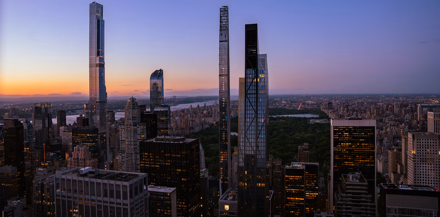 New York City skyline at dusk, featuring tall skyscrapers against a colorful sunset. Central Park is visible, surrounded by illuminated buildings.