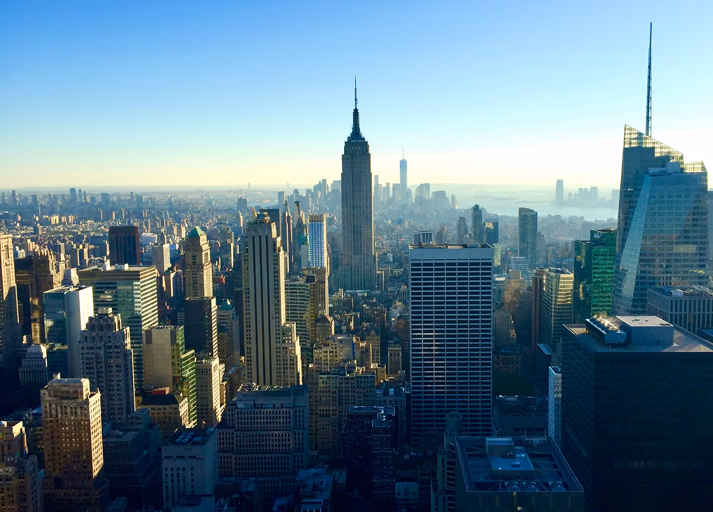 Aerial view of New York City skyline at sunrise, with the Empire State Building prominent. Skyscrapers bathed in golden light, clear blue sky.