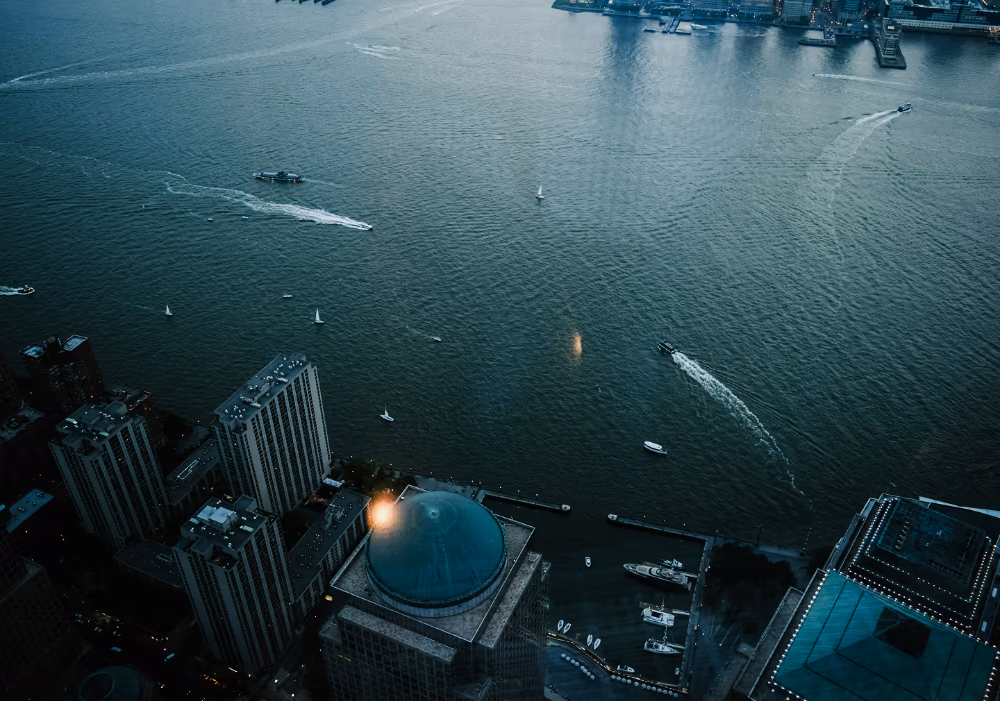Aerial view of a cityscape along a river. Boats leave white trails in the water, and tall buildings with geometric designs are lit by soft evening light.