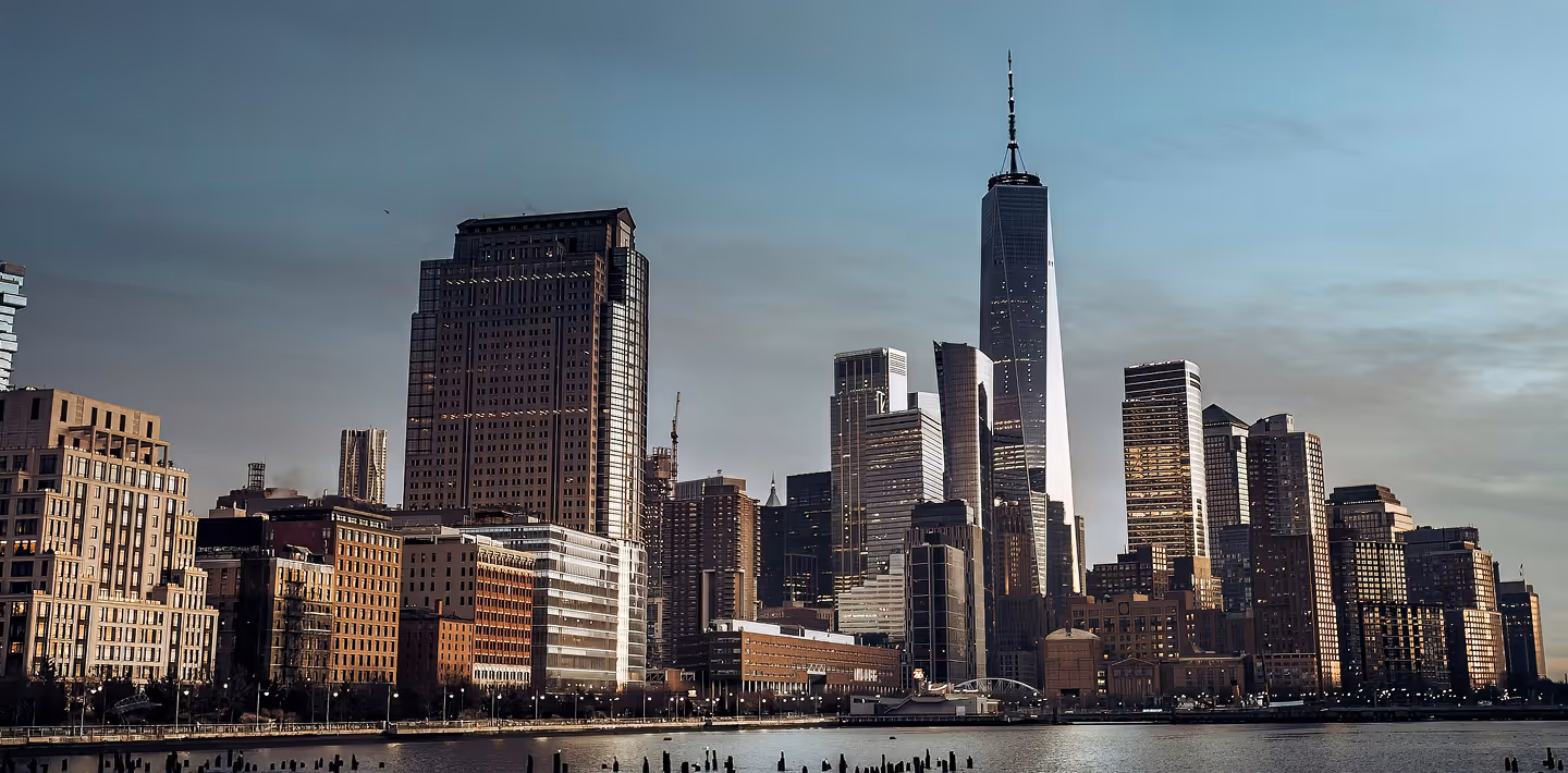 Skyline of New York City at dusk, featuring tall skyscrapers including One World Trade Center. The calm Hudson River is in the foreground, reflecting buildings.