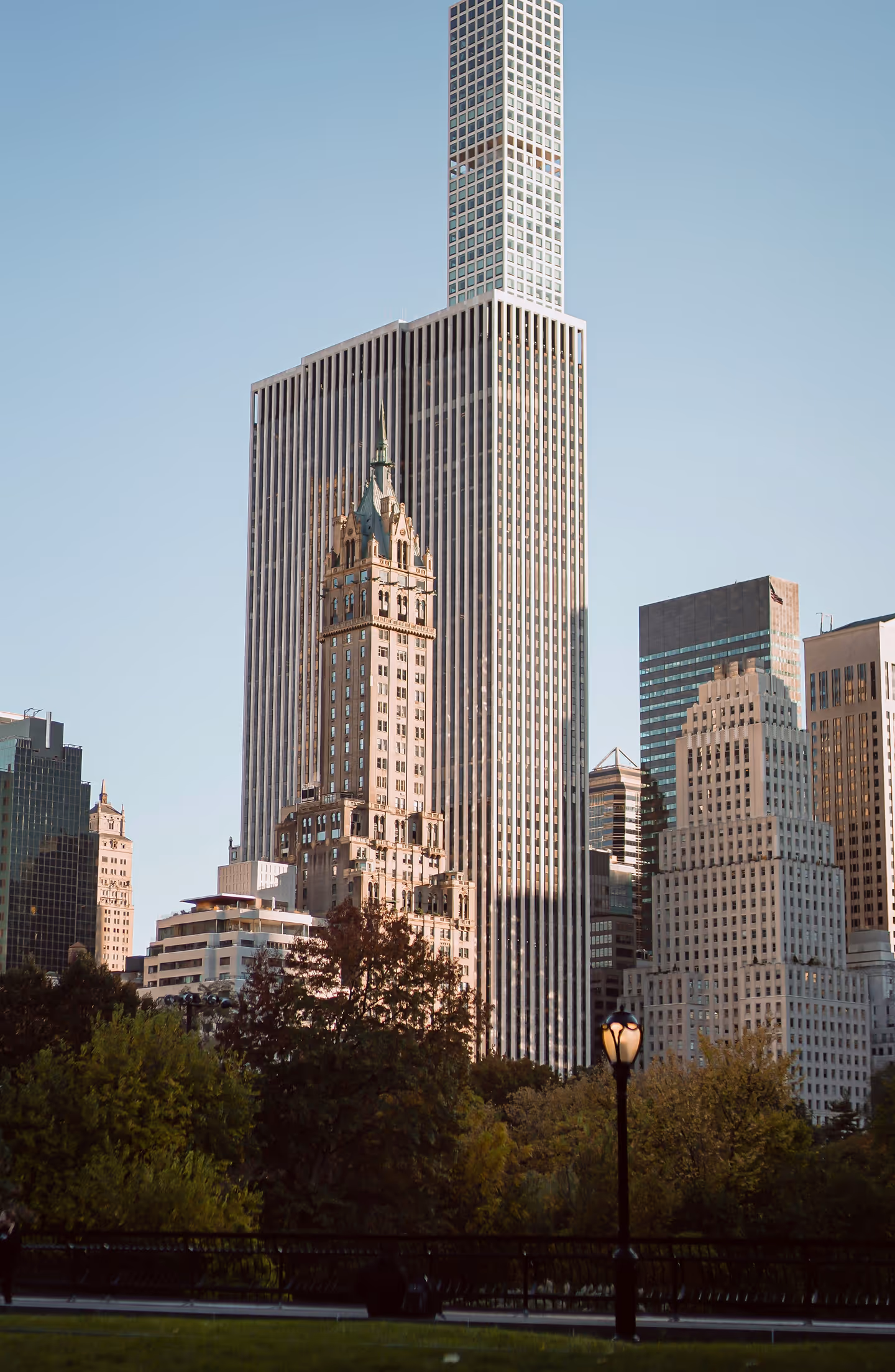 View of a tall, modern skyscraper with a textured facade, surrounded by classic buildings, greenery, and a lamppost under a clear blue sky. Urban and serene scene.