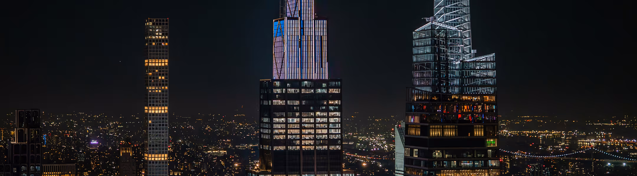 Nighttime cityscape featuring illuminated skyscrapers against a dark sky. The bright windows and lights create a lively urban atmosphere.