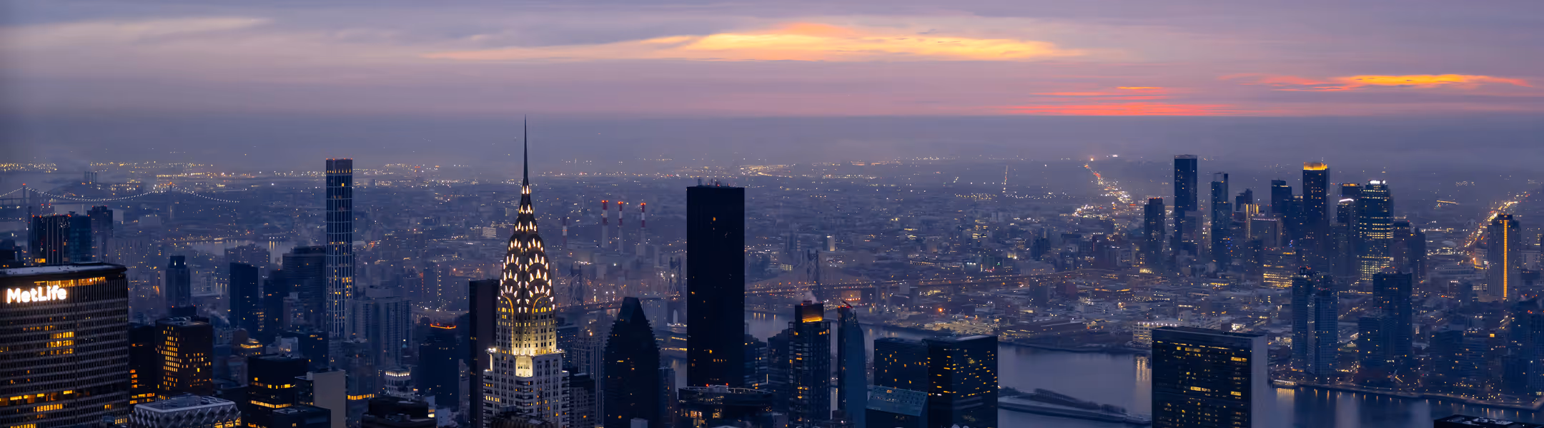City skyline at dusk, featuring lit skyscrapers and the Chrysler Building. A vibrant sunset with orange hues contrasts against a cloudy sky.