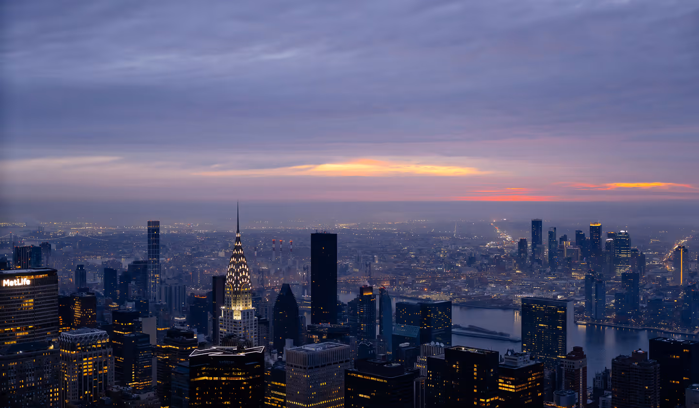City skyline at dusk, featuring lit skyscrapers and the Chrysler Building. A vibrant sunset with orange hues contrasts against a cloudy sky.