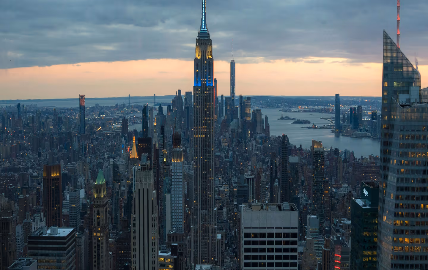 Aerial view of New York City skyline at dawn, with the Empire State Building prominent. Skyscrapers bathed in golden light, clear blue sky.