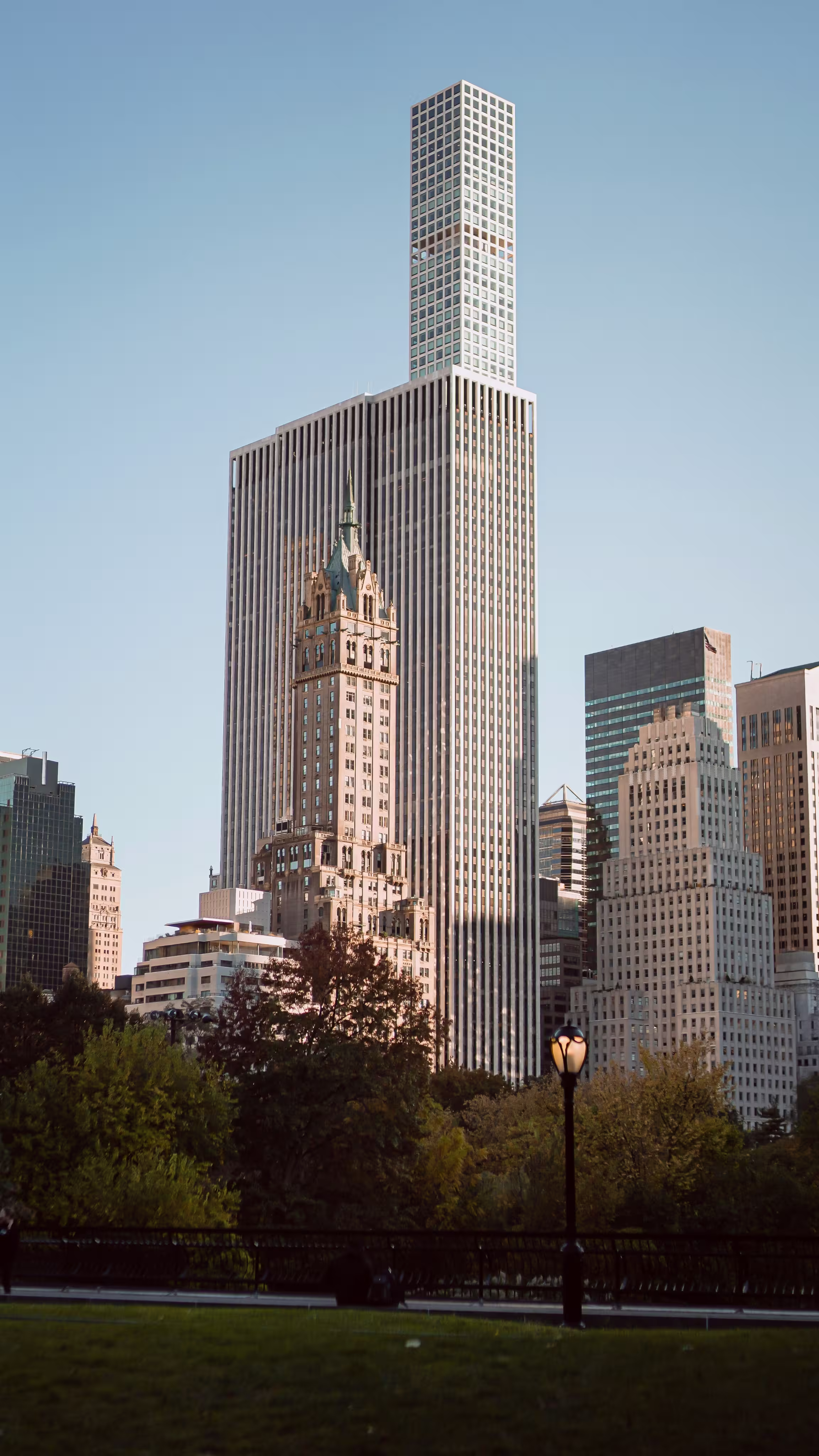 View of a tall, modern skyscraper with a textured facade, surrounded by classic buildings, greenery, and a lamppost under a clear blue sky. Urban and serene scene.