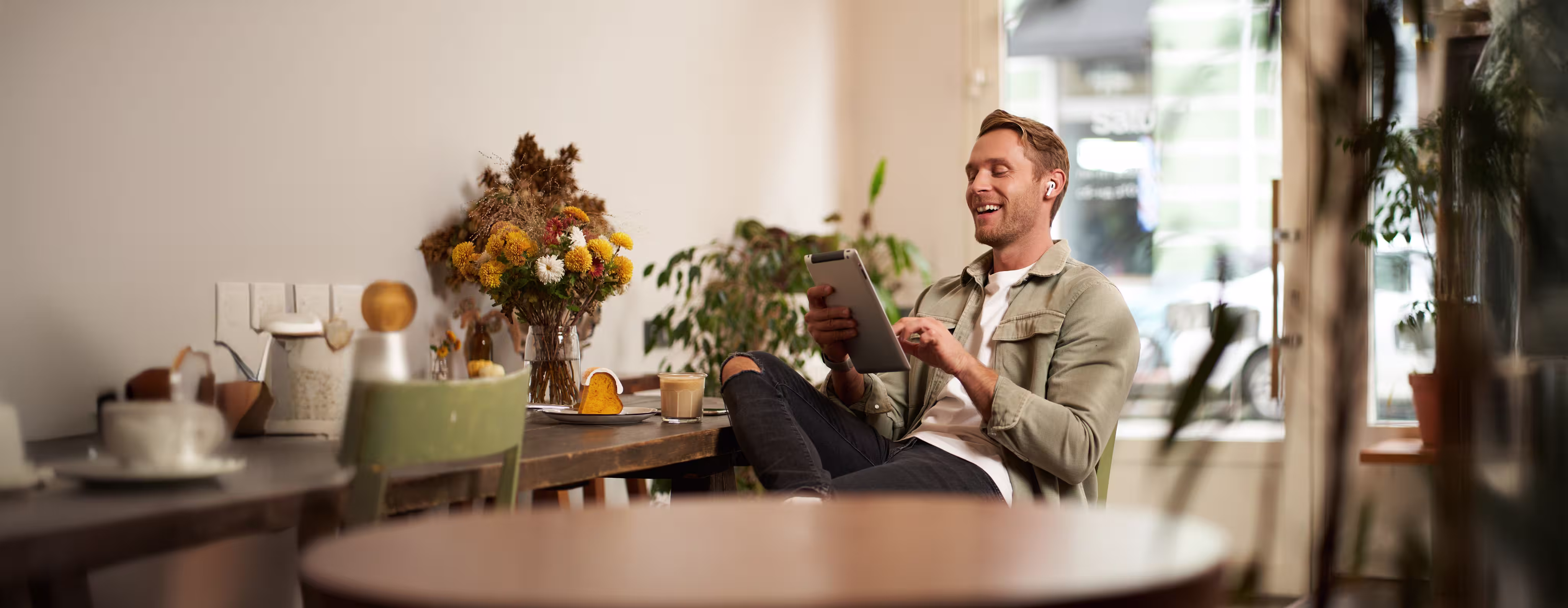 A man in a casual outfit happily sits at a cafe table, holding a tablet. Nearby, a vase with flowers adds a cozy touch. The scene is bright and relaxed.