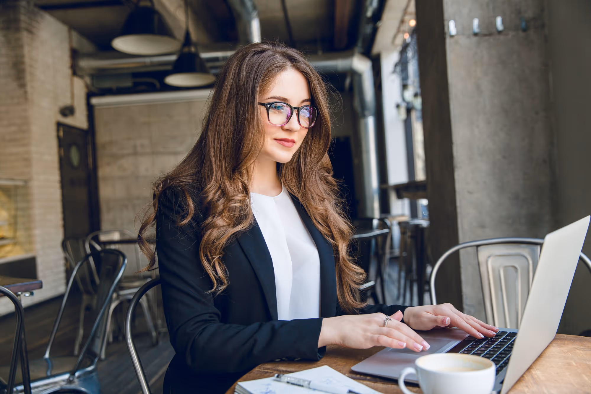 A woman in a black blazer works on a laptop in a modern café, with a coffee cup nearby. She appears focused, conveying a professional and calm atmosphere.