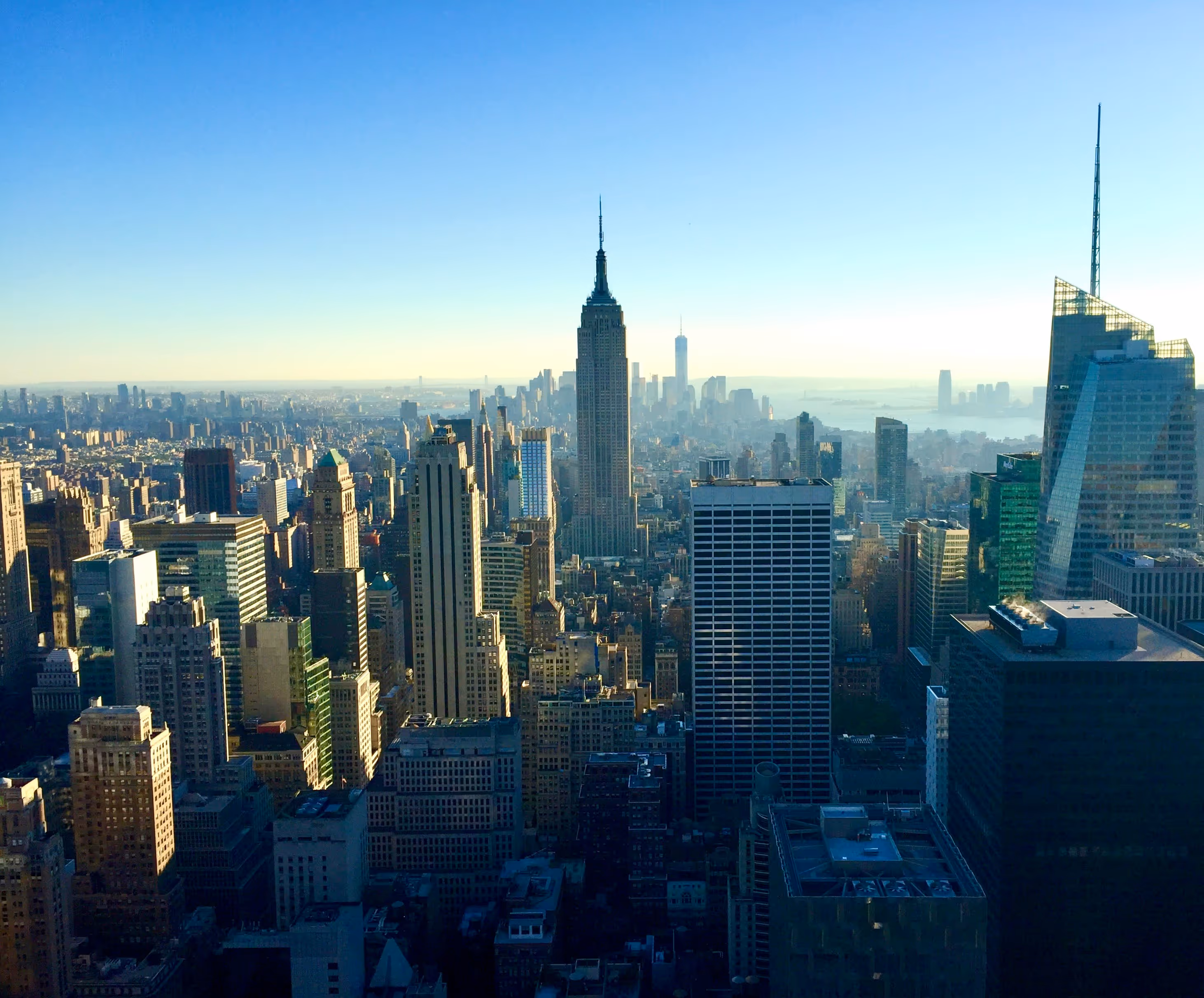 Aerial view of New York City skyline at sunrise, with the Empire State Building prominent. Skyscrapers bathed in golden light, clear blue sky.