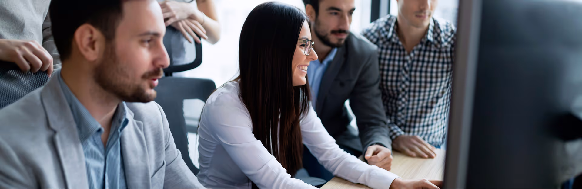 A diverse group of professionals in business attire gather around a computer screen, collaborating with focused, engaged expressions in a bright office setting.