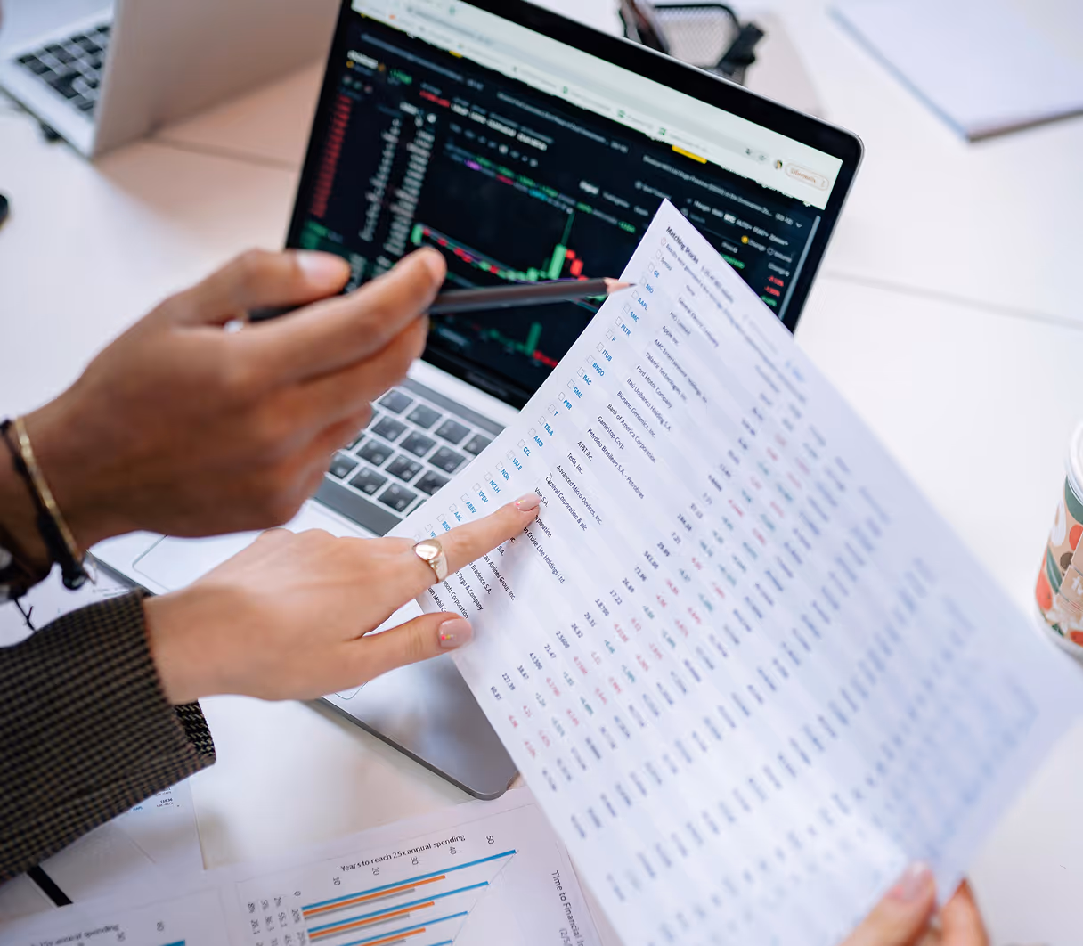 Two people review financial charts and tables at a desk. A laptop displays stock market graphs. The scene conveys analysis and collaboration.