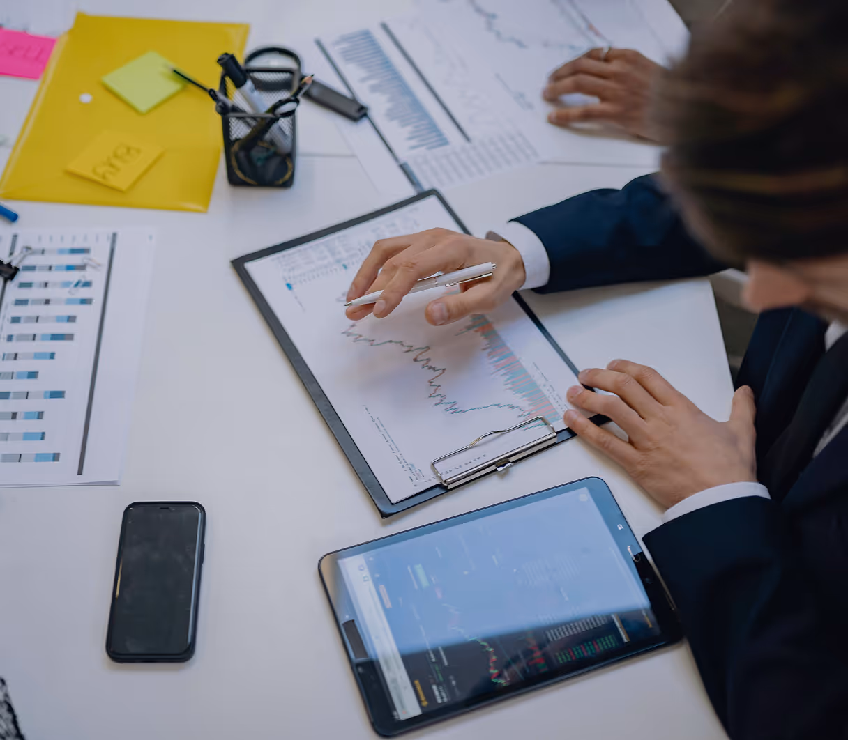 Business professionals analyzing financial graphs on paper and tablets at a desk. A person points to a chart, creating a focused and analytical mood.