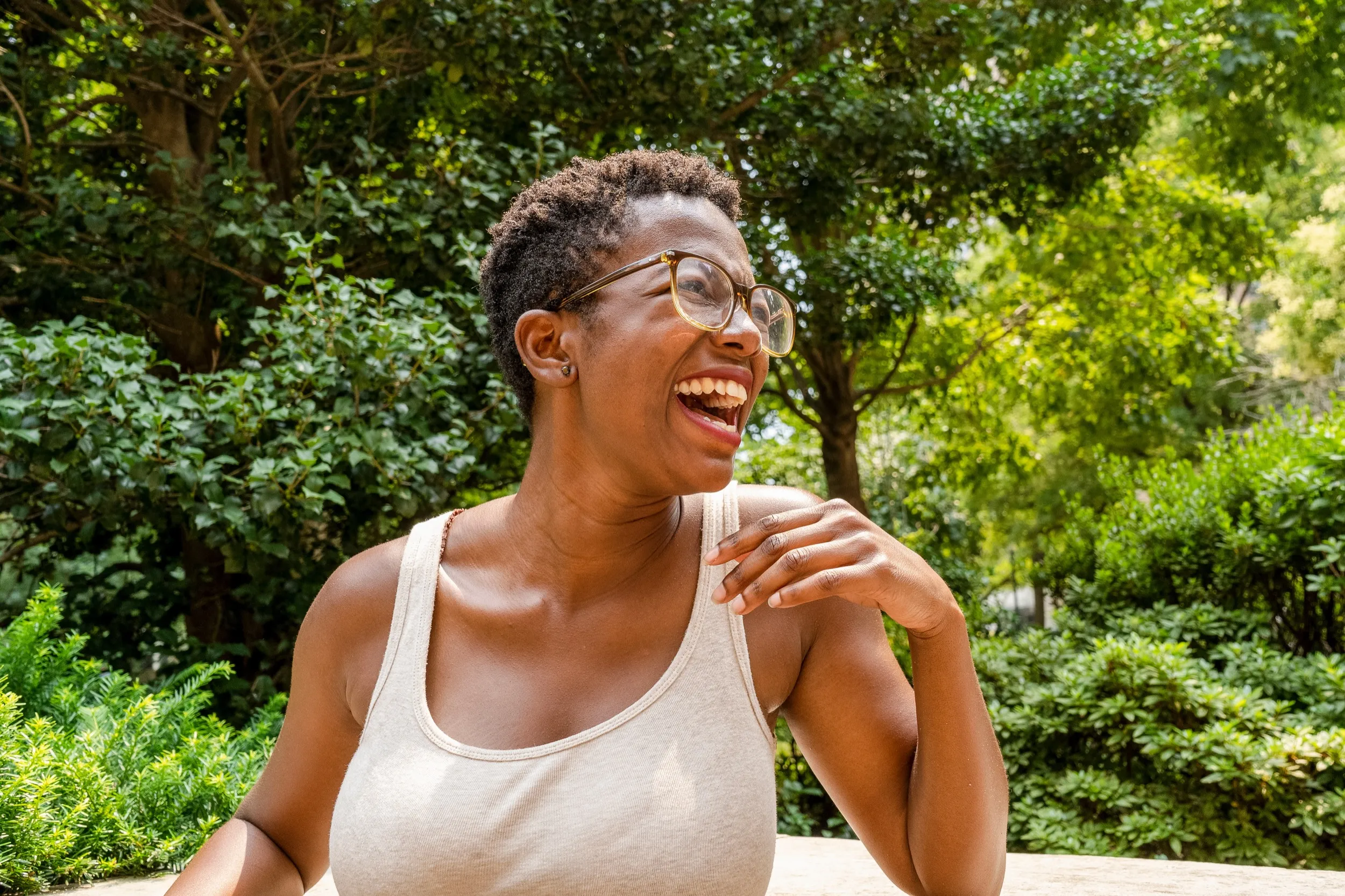 Person in white tank top laughing outdoors among green leafy trees