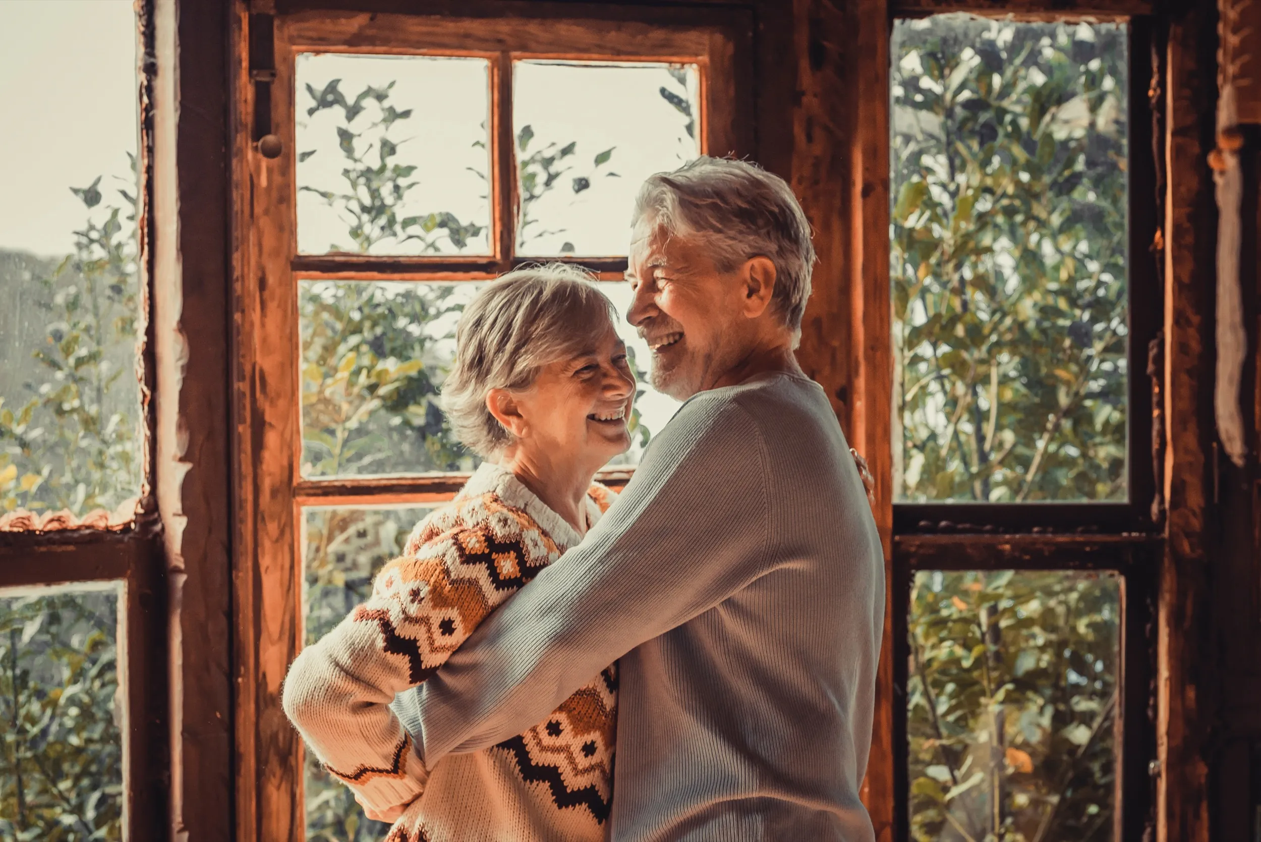 Elderly couple hugging and laughing near window with greenery background