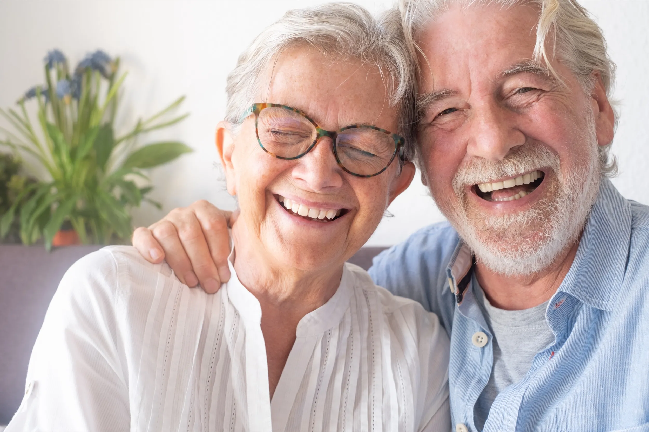 Elderly couple laughing together, wearing glasses, with houseplant in background