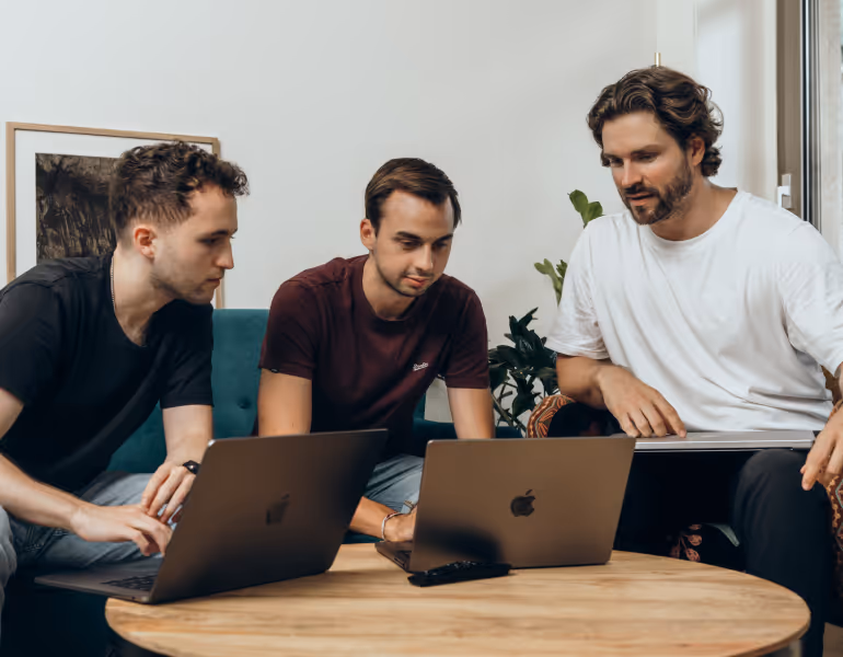 Drei Männer sitzen an einem runden Holztisch und arbeiten gemeinsam an zwei geöffneten MacBook-Laptops.
