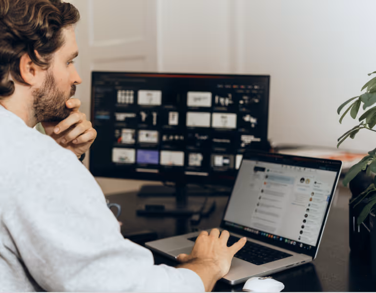 Ein Mann sitzt am Schreibtisch und betrachtet nachdenklich einen Laptop-Bildschirm mit Chat-Interface, im Hintergrund ein größerer Monitor mit verschiedenen geöffneten Fenstern.