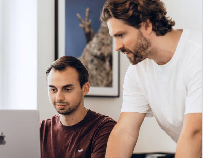Zwei Männer konzentrieren sich auf einen Laptop, während einer sitzt und der andere über ihn gebeugt steht.