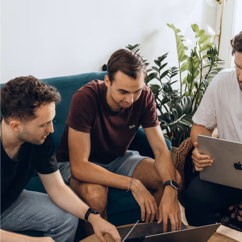 Drei Männer sitzen auf einem Sofa und arbeiten zusammen an Laptops, umgeben von Zimmerpflanzen.
