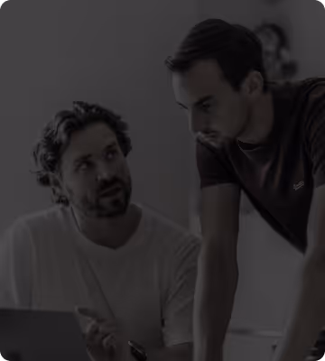 Zwei Männer unterhalten sich konzentriert in einem Büro, einer sitzt und zeigt mit der Hand, der andere steht und hört zu.