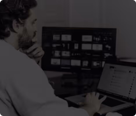 Ein Mann arbeitet konzentriert an einem Laptop mit offenem Chatfenster, während ein Monitor mit mehreren Anwendungsfenstern im Hintergrund zu sehen ist.