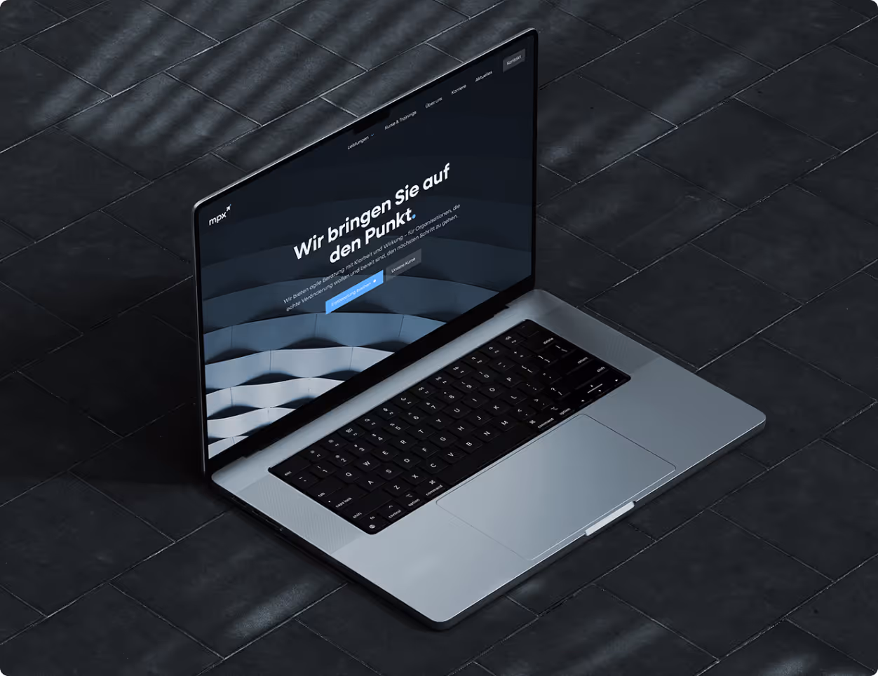 Silberner Laptop auf dunklem Fliesenboden mit geöffneter Webseite, die deutschen Text und Navigationsmenü zeigt.